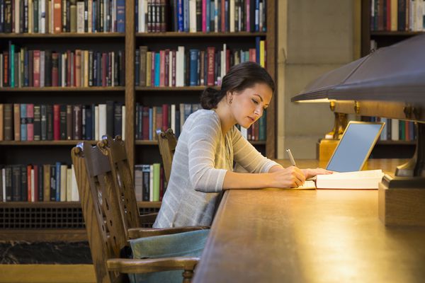 一位女士在图书馆的长木桌上用笔记本电脑写研究论文